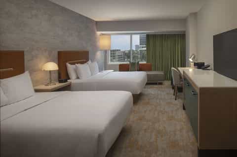 Modern hotel bedroom with two queen beds, work desk, and floor-to-ceiling windows with city views