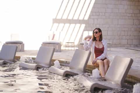 Woman sitting on white lounge chair at beachfront with waves, sand, brick wall backdrop, and sunlight casting geometric shadows