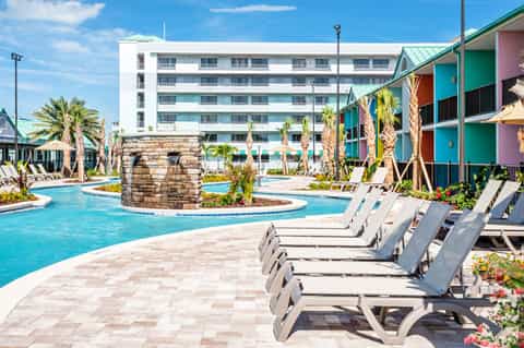 Resort pool area with colorful cabanas, loungers, and multi-story hotel building under blue sky