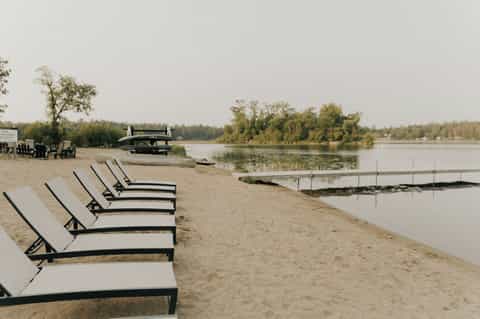 Row of lounge chairs on sandy beach facing calm river with trees beyond