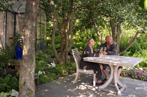 Couple dining outdoors under tall trees with wine glasses at whitewashed table