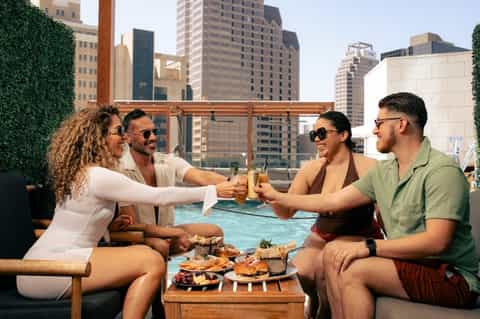 Group of friends toasting by rooftop pool with city skyline and food spread visible