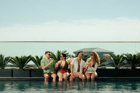 Four people enjoying drinks by rooftop pool with palm trees and coastal view
