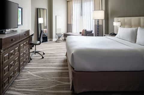 Modern hotel bedroom with king bed, TV, work desk, and floor-to-ceiling windows
