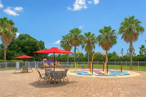 Outdoor playground with red umbrellas, colorful splash pad, and palm trees