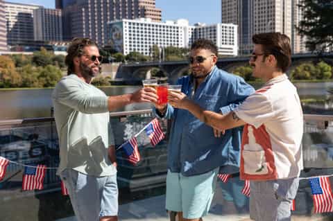 Three men toasting drinks on rooftop terrace with city skyline, river, and American flags