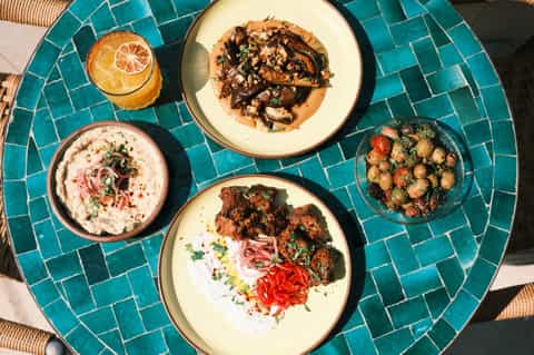 Mediterranean-style appetizers on turquoise tiled table: hummus, grilled vegetables, bread, and fresh juice