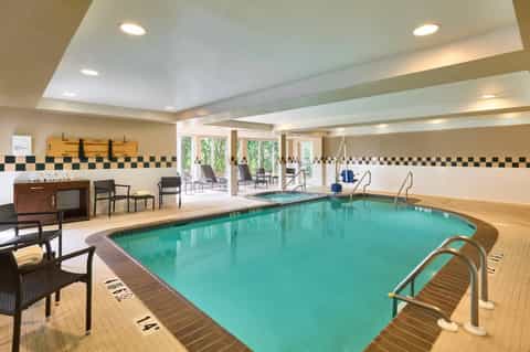 Indoor swimming pool with turquoise water, black and gold tile border, lounge chairs, and large windows overlooking outdoor area