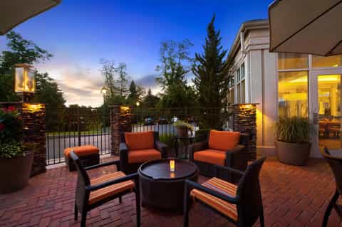 Twilight rooftop patio with wicker seating, orange cushions, fire pit, stone pillars, and lit building facade