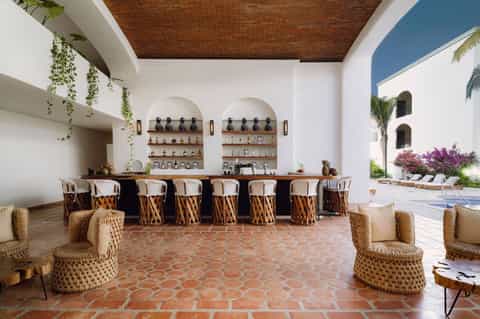 Outdoor bar with woven wicker bar stools, arched shelving displaying bottles, and resort courtyard views.
