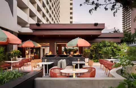 Modern outdoor patio with fireplace, coral umbrellas, woven seating, and tropical plants at dusk