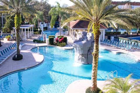 Expansive resort pool complex with palm trees, white cabanas, multiple pools, and lounge chairs
