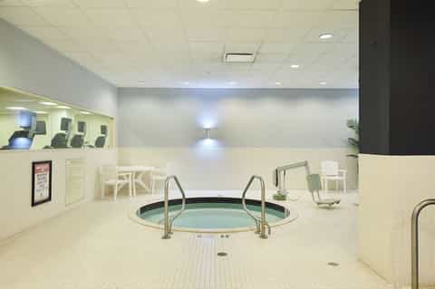 Indoor hot tub with metal railings in a cream tile spa room with fitness equipment visible in background