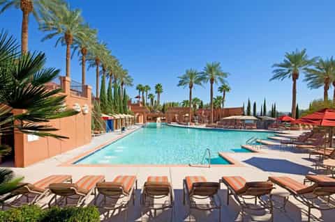 Resort pool with lounge chairs, palm trees, Moroccan-style architecture, and blue umbrellas