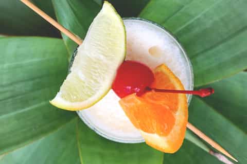 Tropical cocktail with lime wedge, cherry, and orange slice garnish against green palm fronds