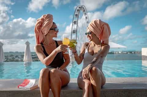 Two women toasting drinks in an infinity pool with Ferris wheel visible in the background