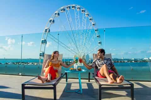 Couple toasting drinks on lounge chairs with a ferris wheel and turquoise bay view