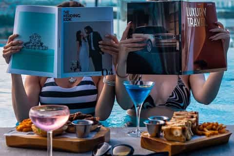 Two women in swimwear holding luxury travel and wedding magazines by a pool with cocktails and appetizers