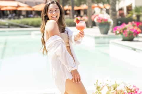 Woman holding cocktail poolside wearing white cover-up with vibrant flowers and resort backdrop