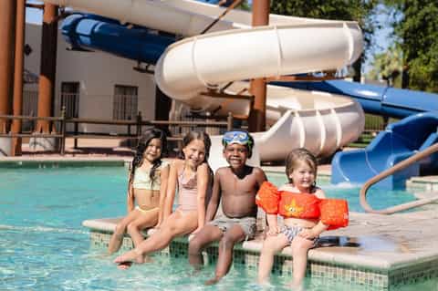 Four children sitting at pool edge with water slides and lazy river in background