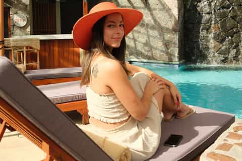 Woman in coral hat relaxing poolside on lounge chair beside turquoise infinity pool