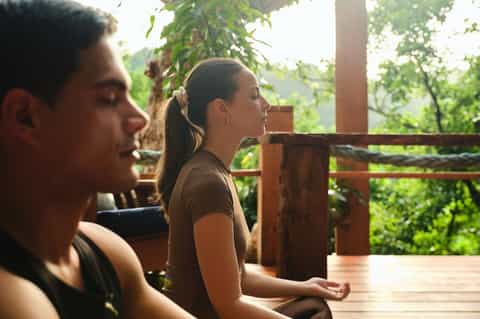 Two people meditating on a wooden deck surrounded by tropical forest greenery
