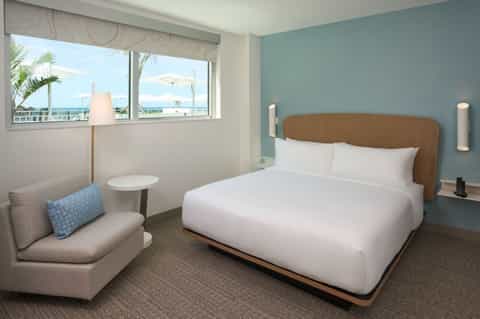 Hotel bedroom with light blue walls, wooden headboard, and window overlooking beach