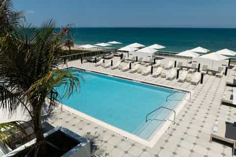 Beachfront resort pool deck with blue water, white umbrellas, lounge chairs, and ocean views