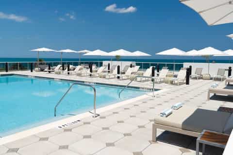 Resort pool area with white umbrellas, lounge chairs, ocean backdrop, and clear blue sky