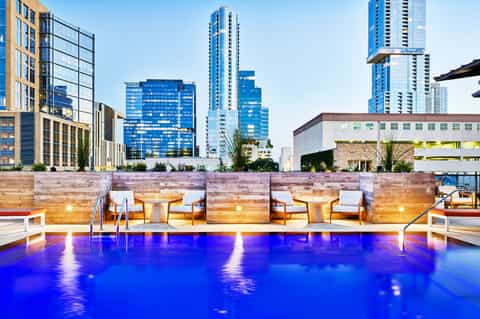 Modern rooftop pool with glowing blue water, wooden loungers, and city skyline backdrop