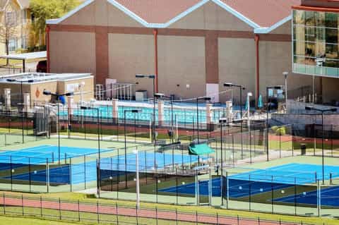 Outdoor tennis courts with blue playing surfaces and protective netting, adjacent to brick facility building