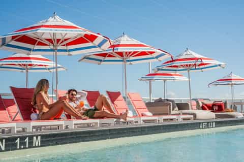 Two guests relaxing in red lounge chairs under red and white striped umbrellas by a shallow pool deck