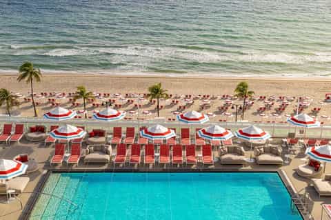 Aerial view of beachfront resort pool, red umbrellas, lounge chairs, and beach with ocean waves