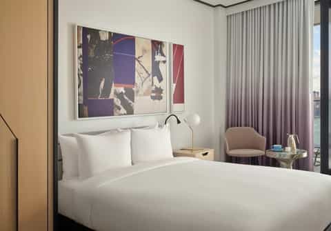 Modern hotel bedroom with white bedding, purple curtains, city skyline view, and wall art