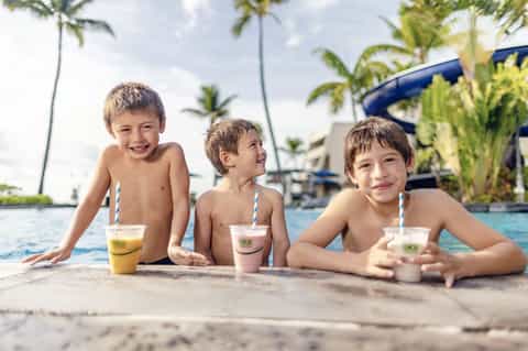Three young children in shallow pool with tropical drinks and palm trees in background