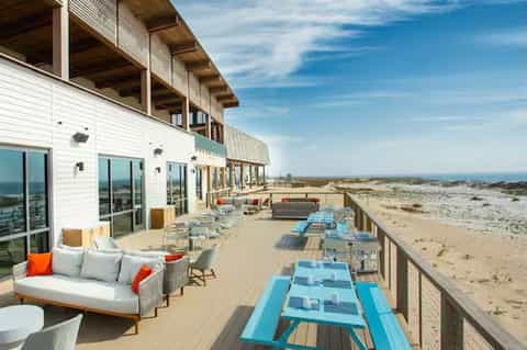 Coastal deck with lounge seating, dining tables, white building, and expansive beach landscape