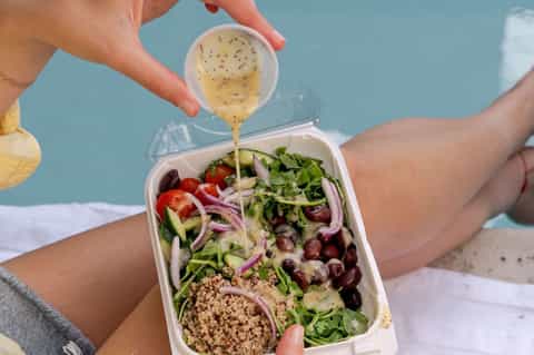 Person drizzling vinaigrette dressing over fresh salad bowl at beach