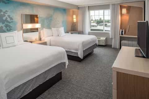 Bright hotel guest room with two queen beds, blue accent wall, river view windows, and modern furnishings