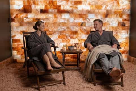 Salt therapy room with two people relaxing in wooden chairs facing illuminated salt brick wall