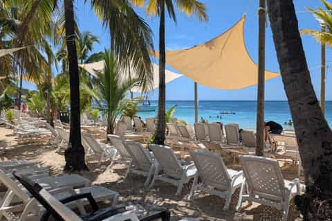 Beach lounge area with white chairs, yellow shade sails, palm trees, and turquoise ocean