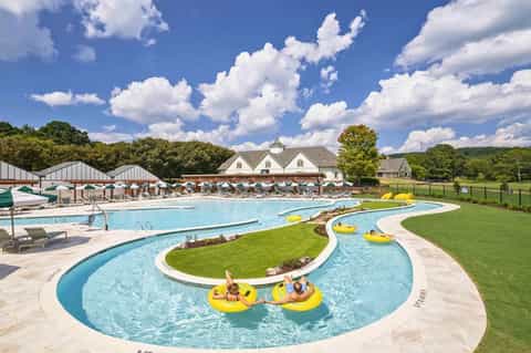 Sprawling resort pool complex with lazy river, yellow floats, lounge chairs, and colonial-style clubhouse under blue sky