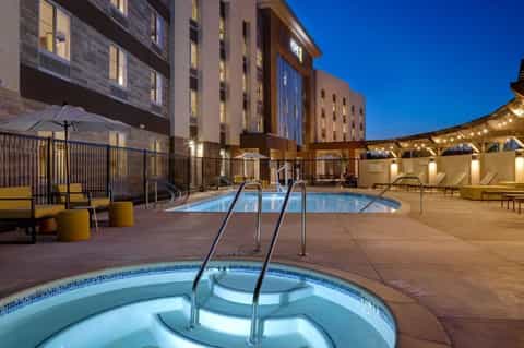 Hotel courtyard at dusk with illuminated hot tub in foreground, swimming pool, lounge seating, and modern brick building facade