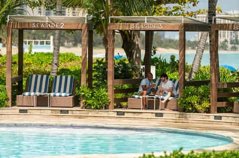 Wooden cabanas with striped cushions beside a resort pool, ocean view in background