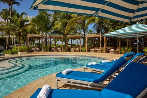 Resort pool with blue loungers, wooden pergolas overhead, striped umbrellas, and palm trees