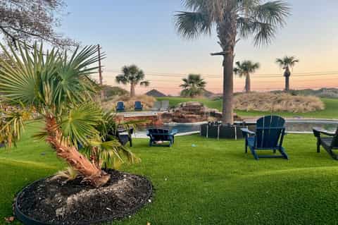 Waterfront property with Adirondack chairs, palm tree, fire pit, dock, and peaceful sunset views