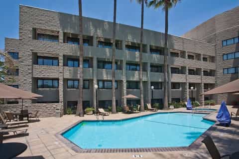Outdoor swimming pool with lounge chairs, stone building facade, palm trees, and desert landscape