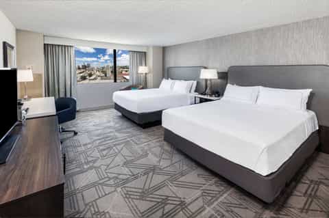 Modern hotel room with two queen beds, city skyline view through windows, work desk, and contemporary décor
