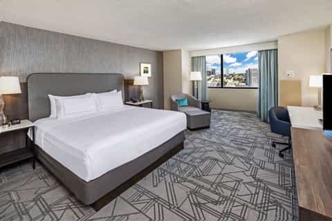 Spacious hotel bedroom with king bed, gray headboard, city view windows, armchair, and geometric patterned carpet