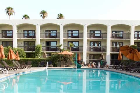 Rectangular pool with orange umbrellas and loungers facing white arched hotel buildings with palm trees