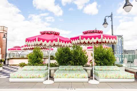 Three ornate pink and cream canopied daybeds with floral cushions on an urban rooftop, framed by green topiaries and city skyline.